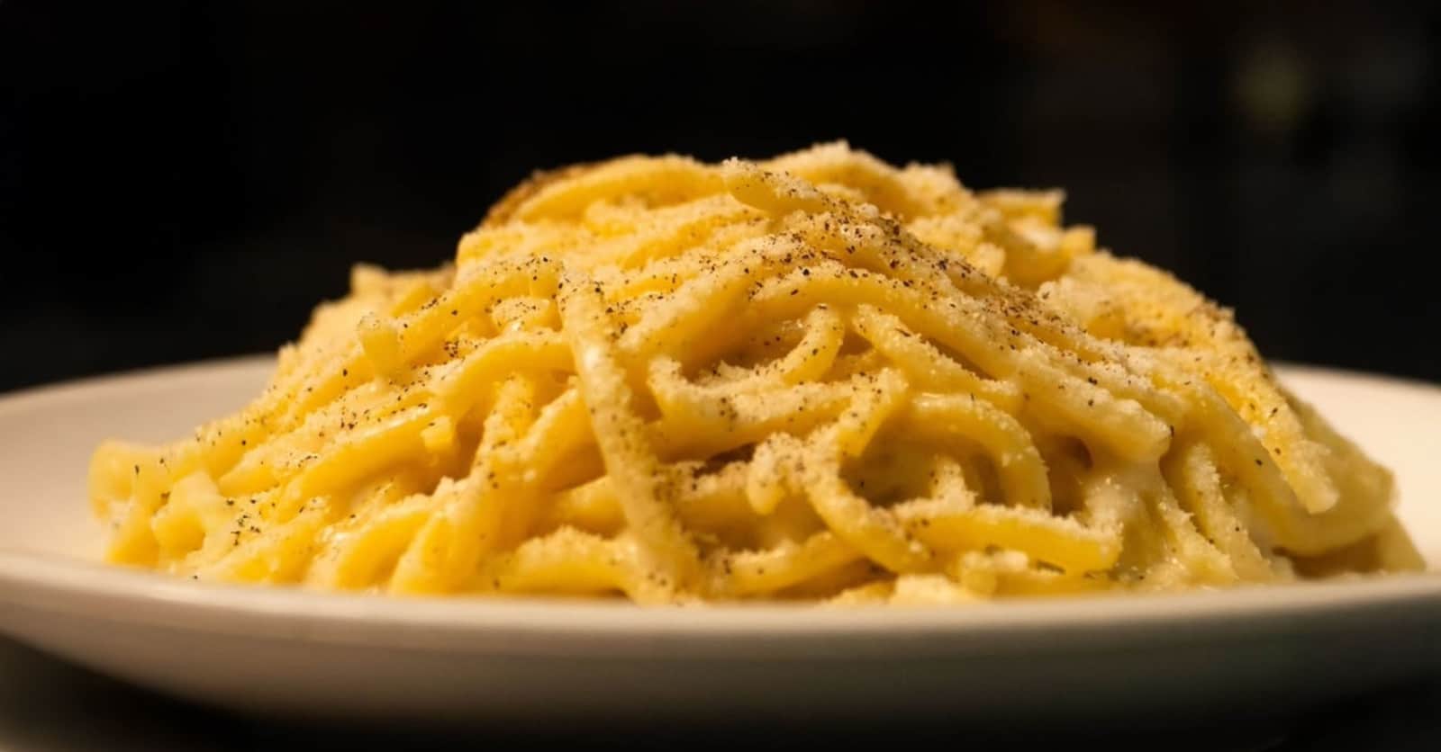 Un cumulo di spaghetti conditi con crema di formaggio e abbondante pepe nero, su piatto bianco e sfondo scuro.