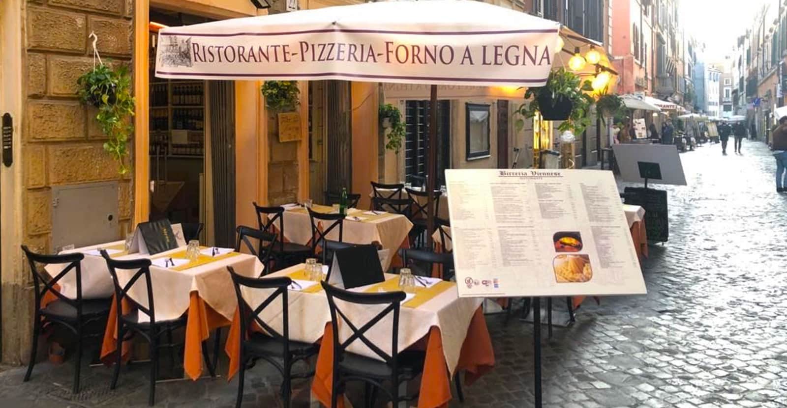 Tavoli con tovaglie bianche e tovaglioli arancioni sotto un grande ombrellone con scritta Ristorante-Pizzeria-Forno a Legna.