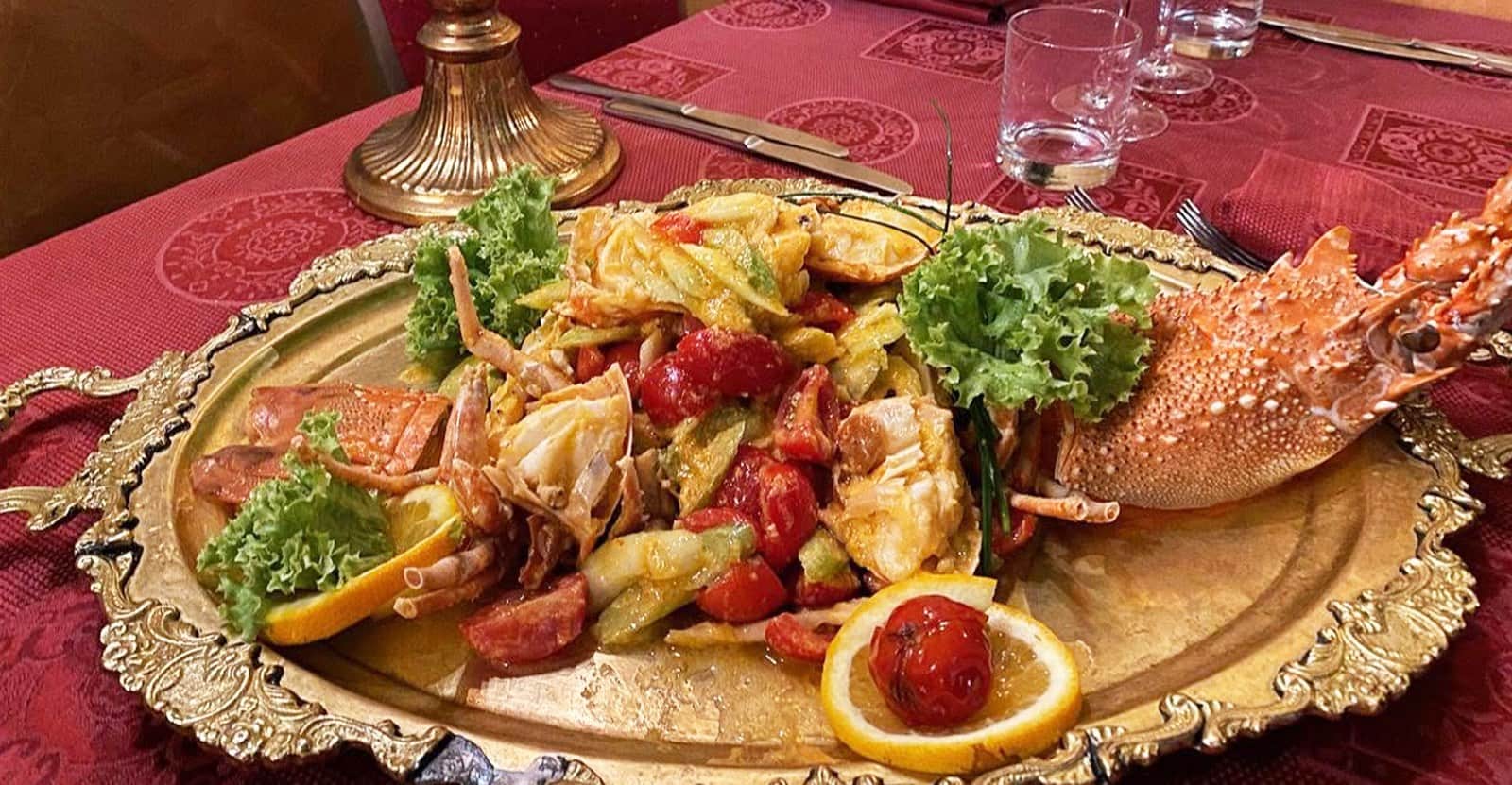 Vassoio dorato con astice, pezzi di granchio, pomodorini e zucchine, guarnito con insalata riccia e fette di limone.