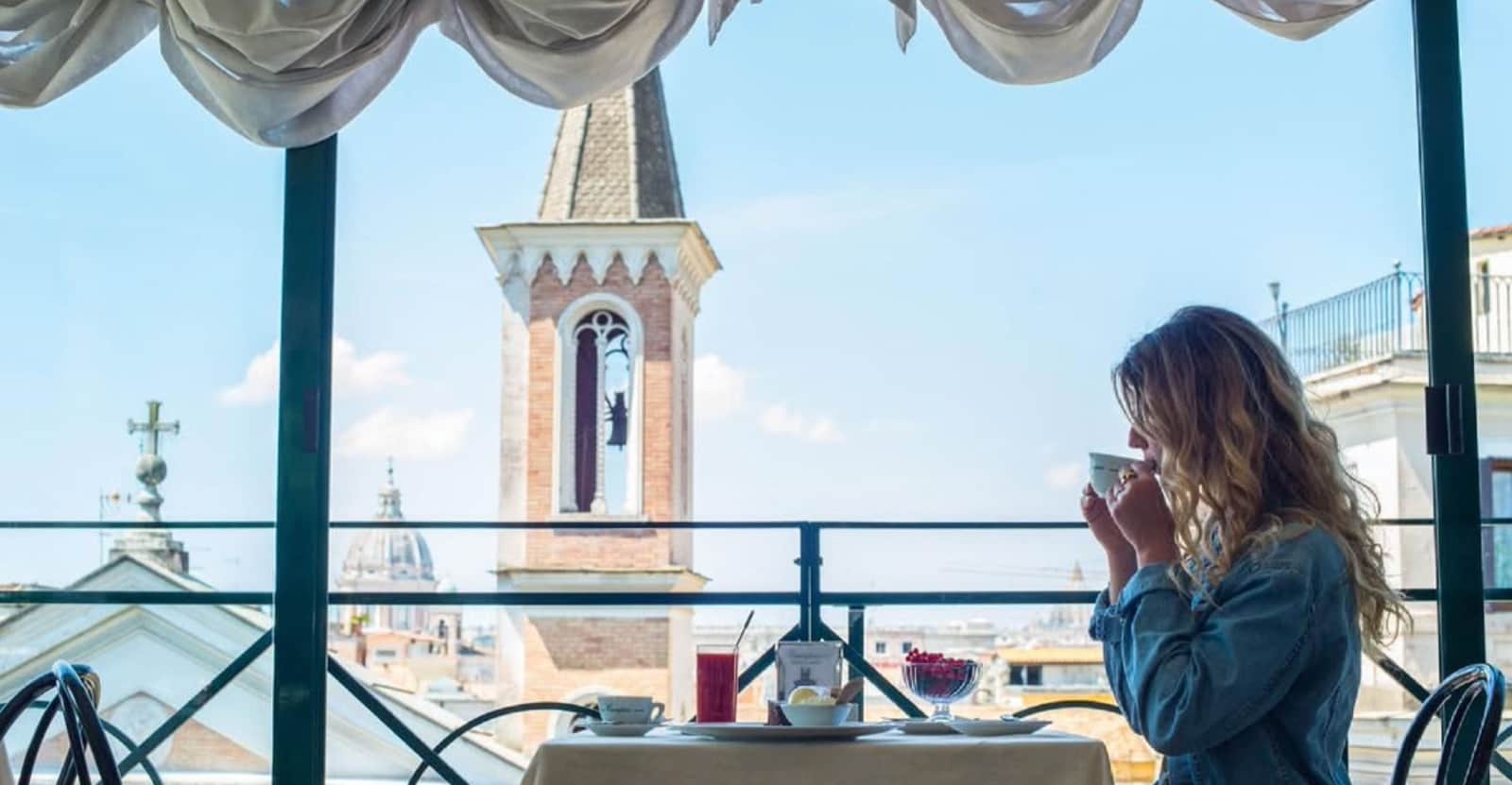 Donna beve da una tazzina su una terrazza, sullo sfondo un campanile in mattoni e il profilo di Roma.