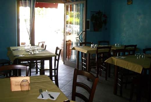 Sala con pareti azzurre, tavoli con tovaglie verde oliva e sedie in legno scuro, porta a vetri aperta sul fondo.