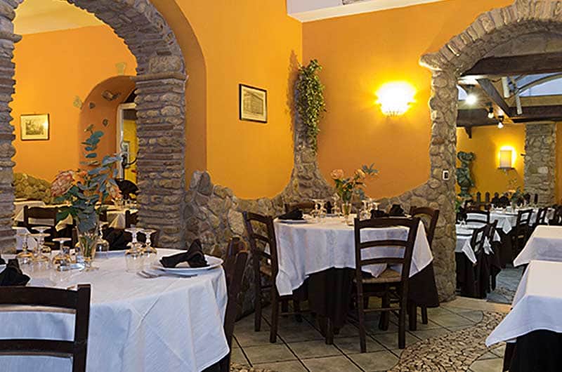 Sala con archi in pietra, pareti color ocra e tavoli apparecchiati con tovaglie bianche e tovaglioli neri.