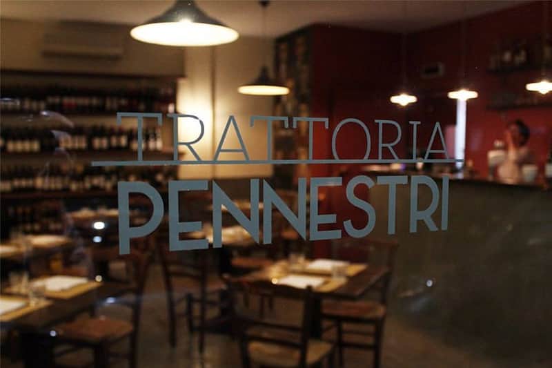 Scritta 'Trattoria Pennestri' su una vetrina, con tavoli apparecchiati e scaffali di vino visibili all'interno.