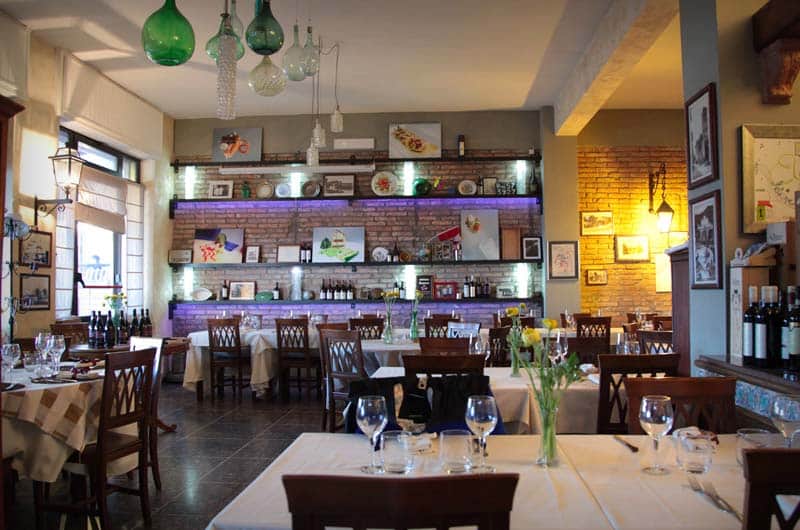 Sala da pranzo con tavoli apparecchiati di bianco, mensole in muratura con piatti e bottiglie, lampade a sospensione in vetro colorato.