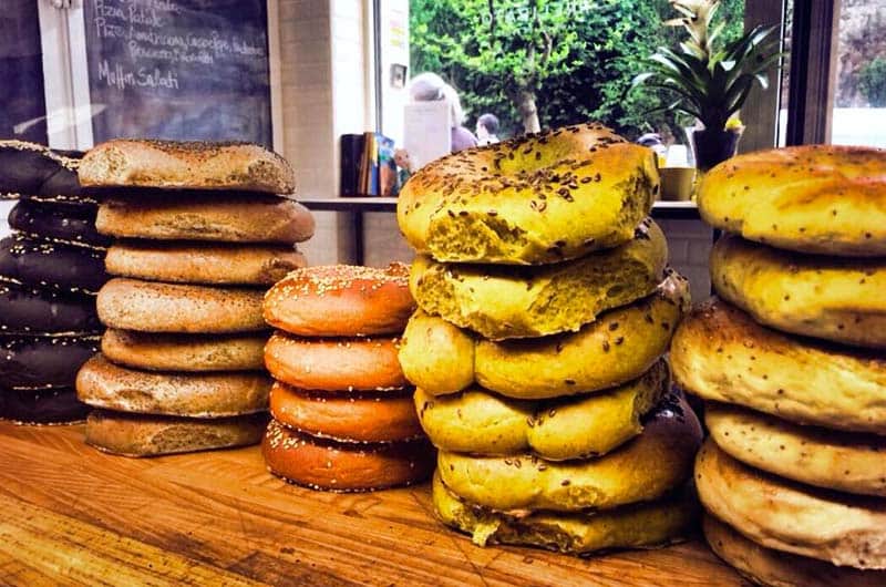Cataste di ciambelle di pane di diversi colori allineate su un bancone di legno, davanti a una vetrata.