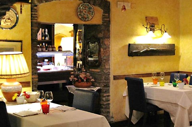 Sala di un ristorante con pareti gialle, tavoli apparecchiati di bianco e bicchieri colorati, separata da un arco in pietra.