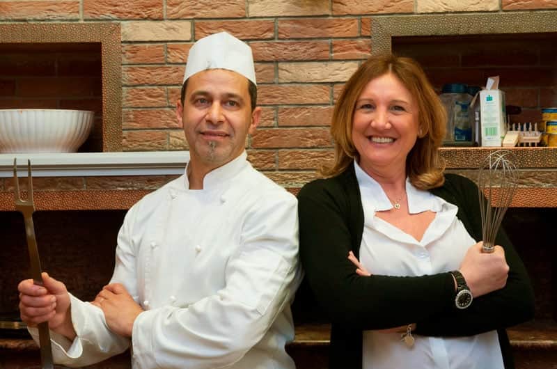 Un cuoco in giacca bianca e cappello da chef e una donna con frusta da cucina in mano, davanti a una parete di mattoni.