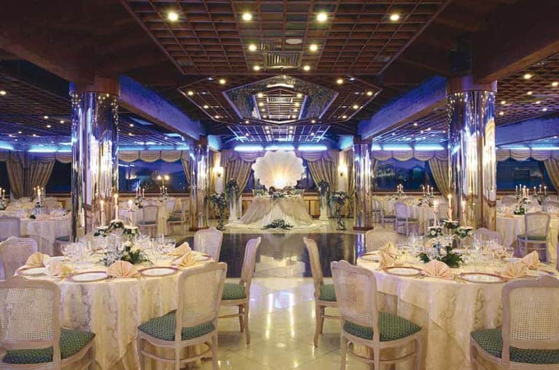 Sala da banchetti con tavoli rotondi apparecchiati, colonne a specchio e luci blu, pista da ballo centrale con composizione floreale bianca.