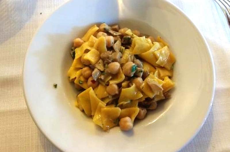 Pasta larga condita con ceci, funghi a dadini ed erbe tritate, servita in un piatto fondo bianco.