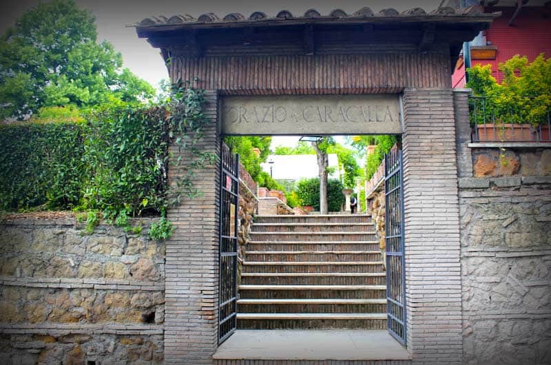 Cancello in ferro battuto aperto su una scalinata di pietra tra due pilastri in mattoni con tetto a tegole.