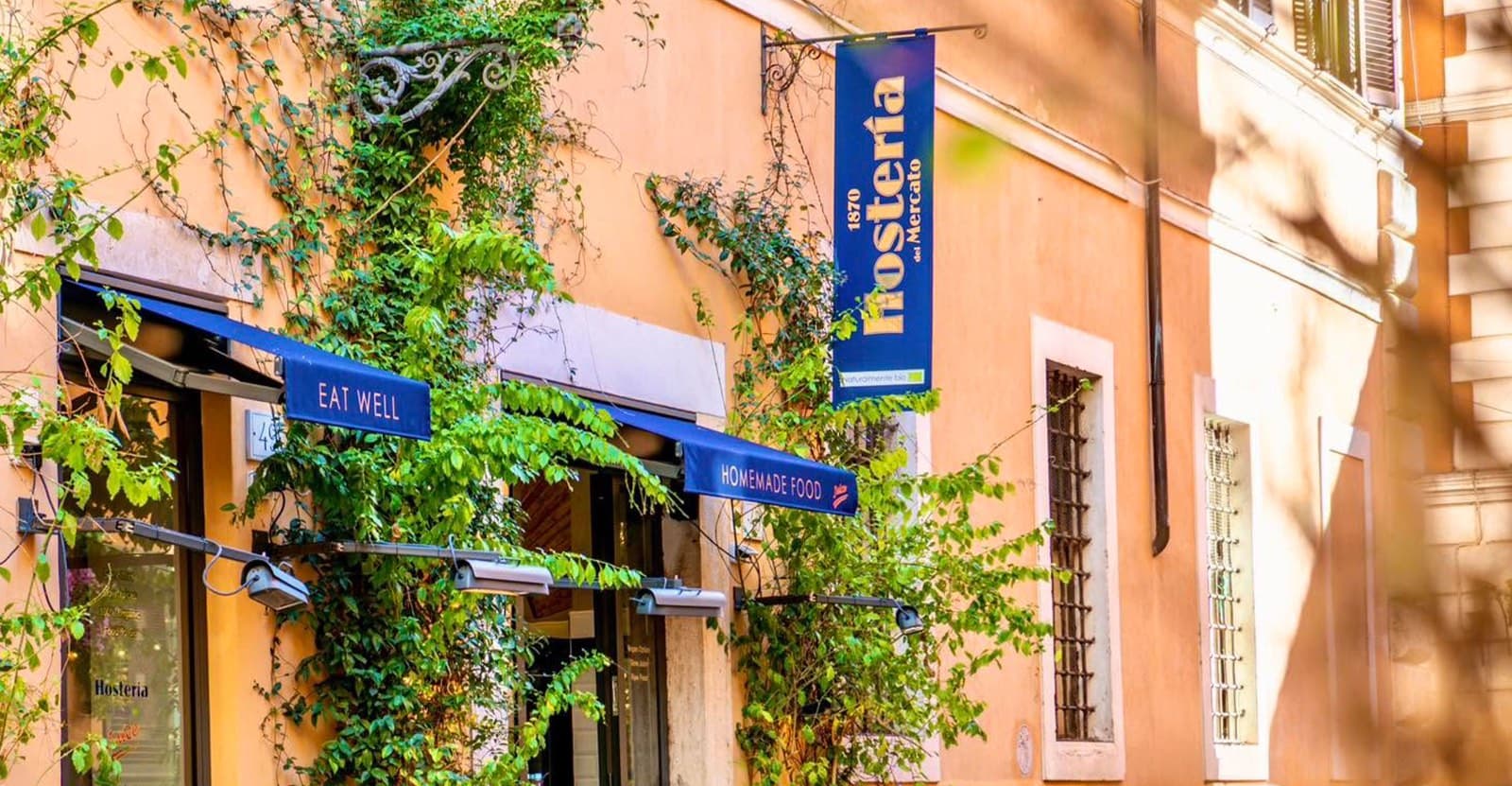 Facciata color ocra con tende blu, una insegna verticale con scritta Hosteria e piante rampicanti sul muro.