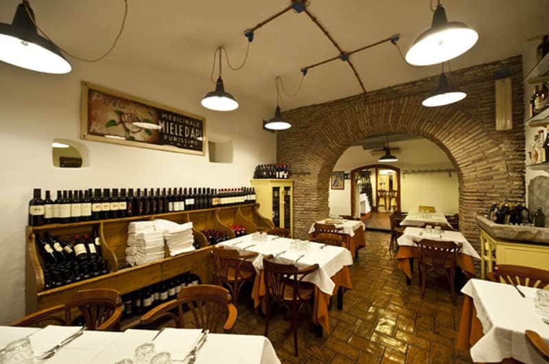 Sala da pranzo con scaffali di bottiglie di vino a muro, tavoli con tovaglie bianche e volta in mattoni sullo sfondo.