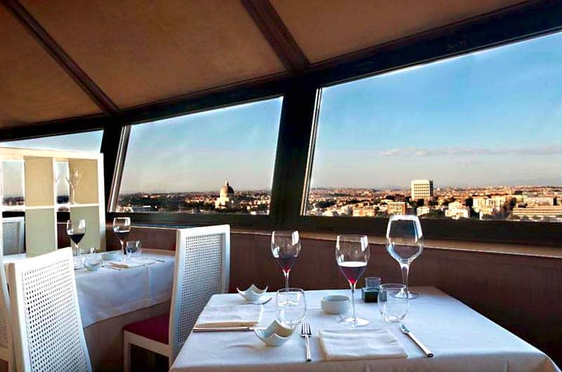 Tavoli apparecchiati con calici da vino accanto a grandi finestre panoramiche sulla città.
