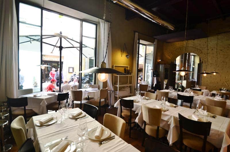 Sala da pranzo con tavoli apparecchiati in bianco, muro in mattoni a vista e lampade a sospensione dal soffitto a travi.