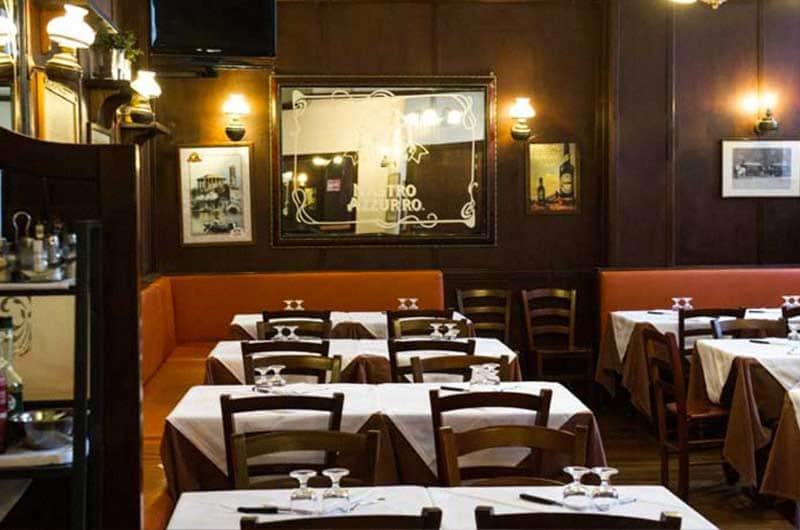 Sala di ristorante con pareti scure, divanetti color ruggine e tavoli apparecchiati con tovaglie bianche e bicchieri da vino.