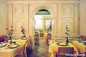 Sala con pareti chiare a rilievo, tavoli quadrati con tovaglie gialle e un arco centrale che conduce a una seconda stanza illuminata.