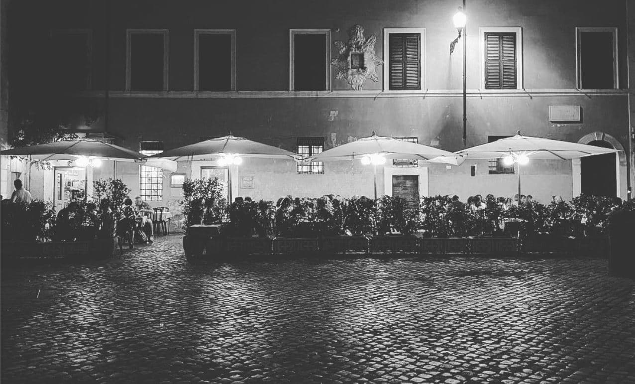 Tavoli all'aperto sotto ombrelloni illuminati, su una piazza di sanpietrini, davanti a una facciata con finestre e persiane.