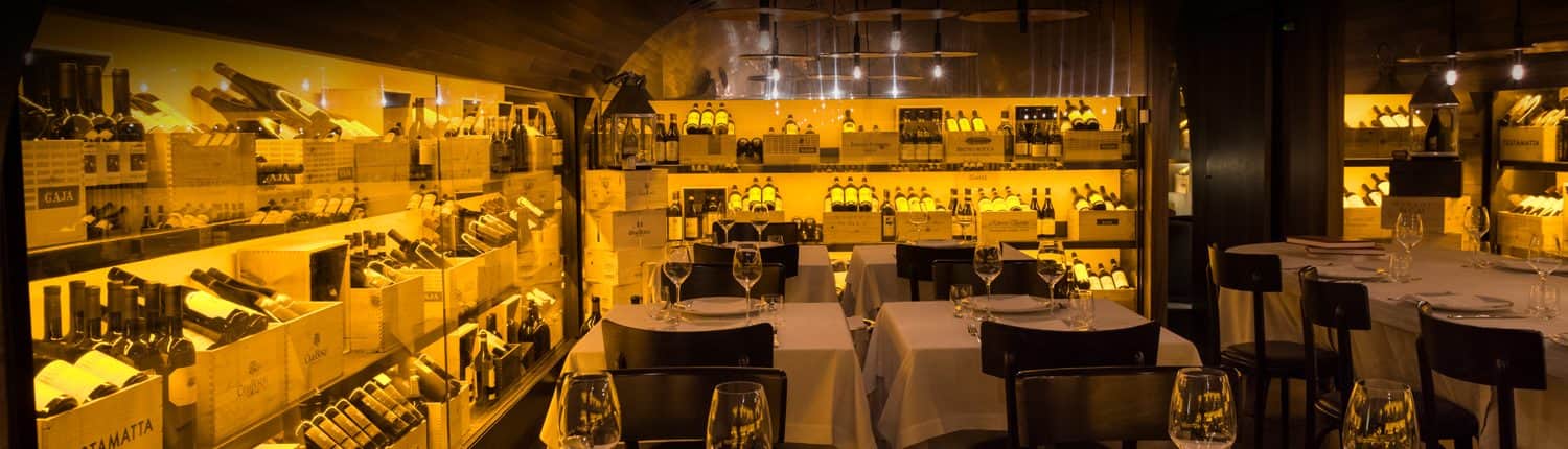 Sala da pranzo con pareti di scaffali illuminati pieni di bottiglie di vino e tavoli apparecchiati con tovaglie bianche.