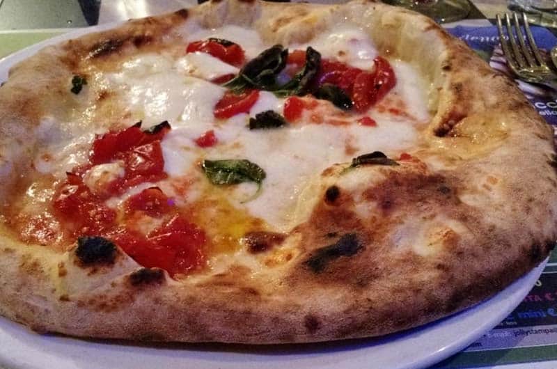 Pizza tonda con crosta alveolata e macchie di cottura scura, farcita con mozzarella filante, pomodori a pezzi e foglie di basilico.