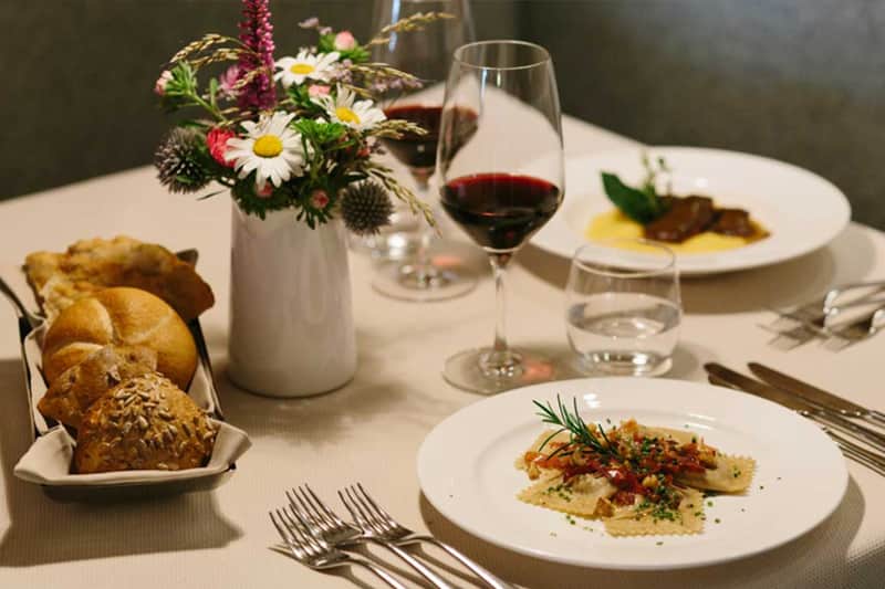 Piatto di pasta ripiena con rosmarino in primo piano, cestino di pane e vaso di fiori di campo sul tavolo.