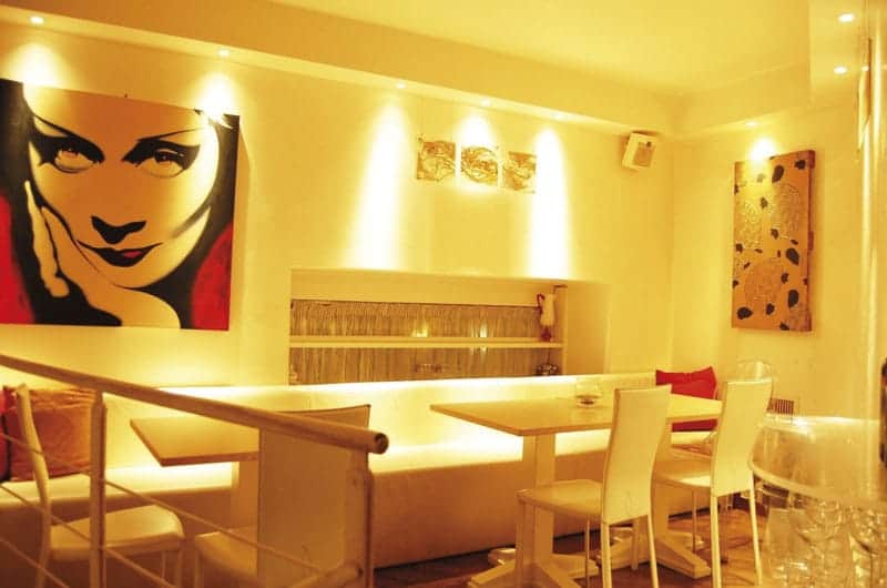 Sala da pranzo con divano bianco, tavoli quadrati e ritratto di donna stilizzato su una parete gialla.
