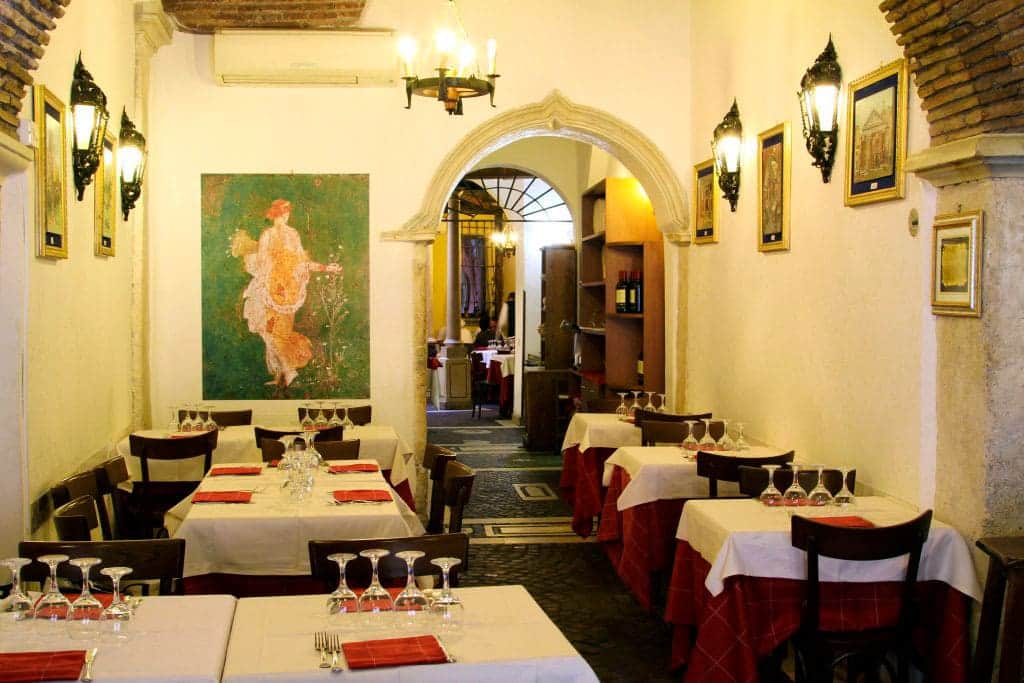 Sala da pranzo con tavoli apparecchiati in bianco e rosso, quadro di una figura femminile e arco in pietra sullo sfondo.