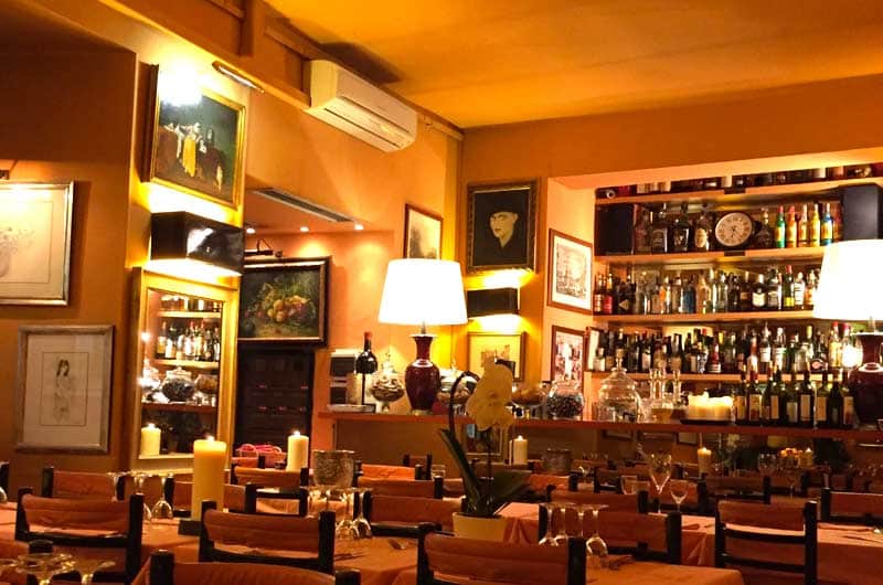 Sala di ristorante con luce calda, tavoli apparecchiati, lampade a paralume bianco e bancone bar con bottiglie sullo sfondo.