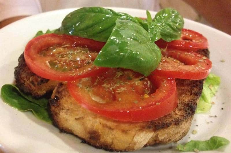 Fetta di pane abbrustolito con fette di pomodoro, foglie di basilico e lattuga su piatto bianco.