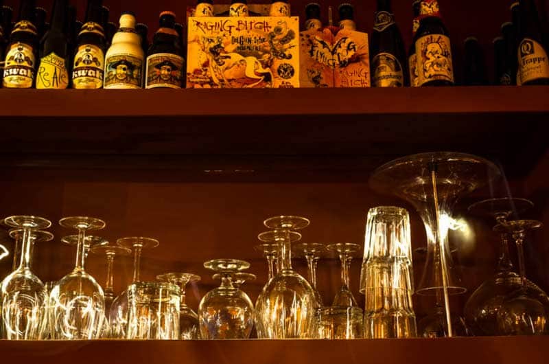 Bottiglie di birra su una mensola sopra bicchieri da vino capovolti illuminati da luce calda.