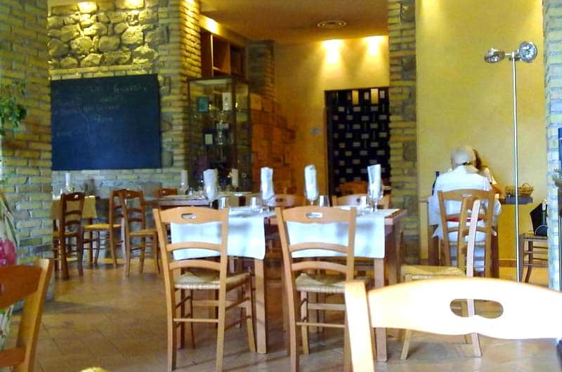 Sala da ristorante con tavoli apparecchiati in bianco, sedie in legno chiaro e pareti in pietra a vista.