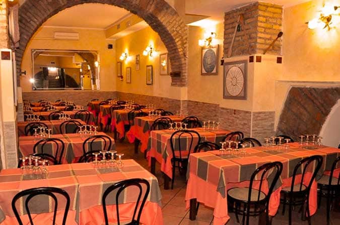 Sala con arco in mattoni, tavoli apparecchiati con tovaglie arancioni e a quadretti, sedie nere in legno curvato.