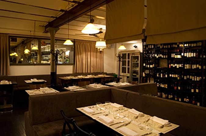 Sala da ristorante con divanetti scuri, tavoli apparecchiati e parete di bottiglie di vino sulla destra.