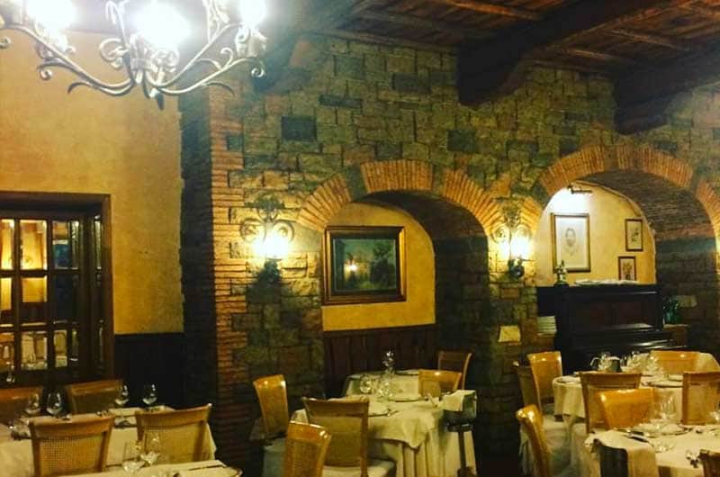 Sala con pareti in mattoni ad arco, tavoli apparecchiati di bianco e un lampadario in ferro battuto al centro.