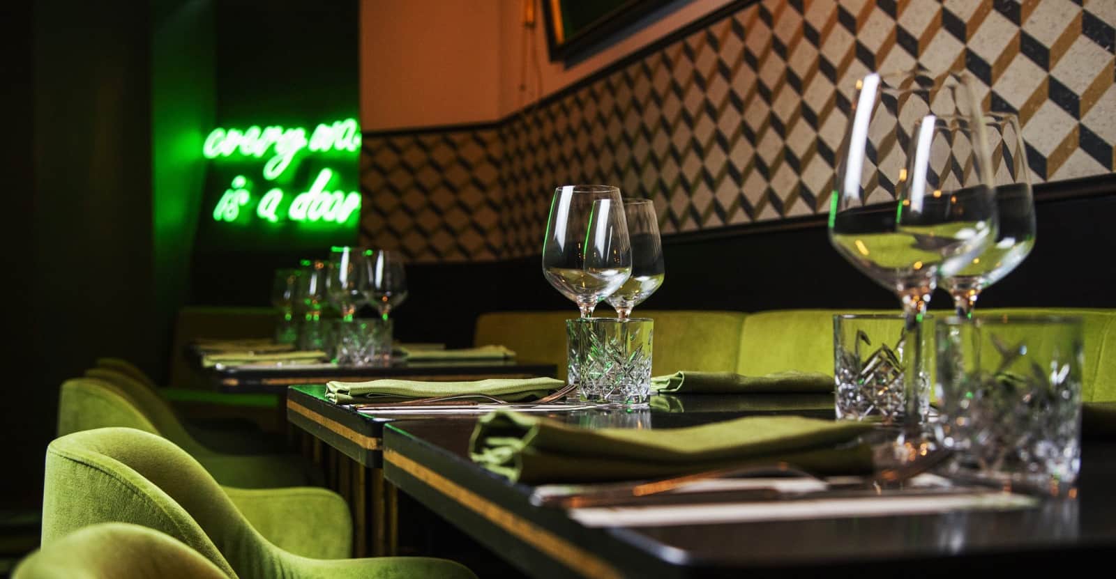 Tavoli apparecchiati con bicchieri di cristallo e tovaglioli verdi, poltrone in velluto verde e insegna al neon sullo sfondo.