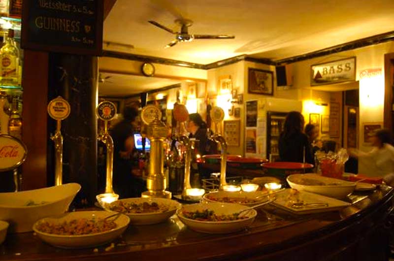 Bancone di bar con ciotole di antipasti in fila, rubinetti dorati della birra e una lavagna con i prezzi delle birre.
