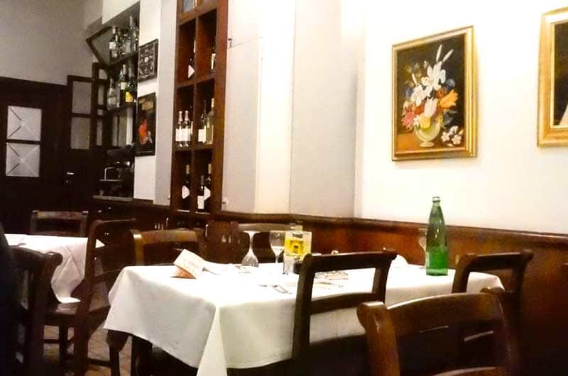 Sala di un ristorante con tavoli apparecchiati di bianco e una libreria di legno scuro con bottiglie sullo sfondo.