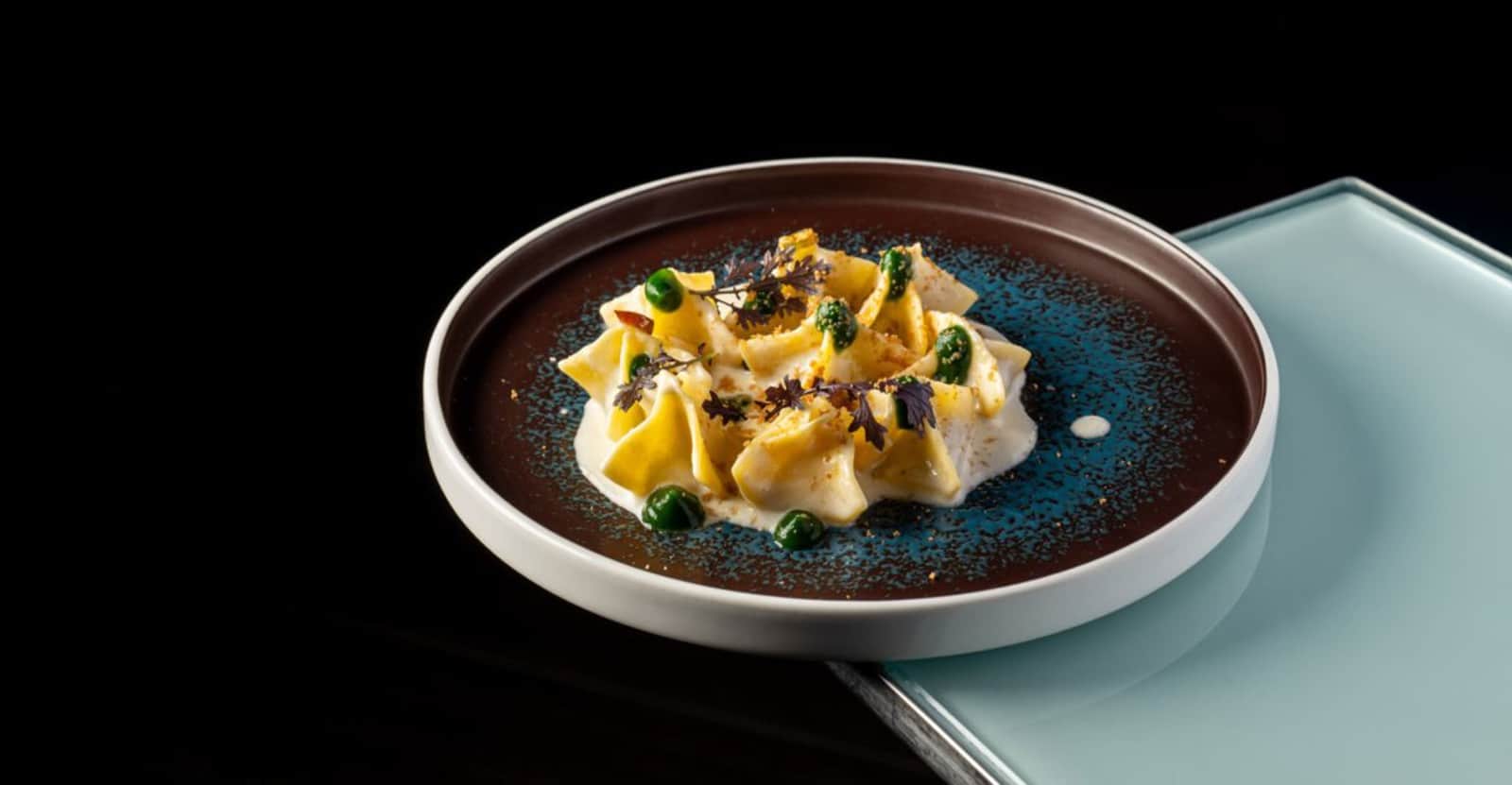 Fazzoletti di pasta gialla su una crema bianca, con piselli verdi ed erbe su piatto scuro con smalto blu.