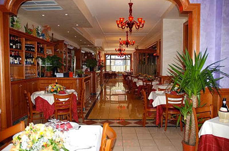 Sala di ristorante con tavoli bianchi e rossi, due lampadari rossi e un bar in legno sulla sinistra.