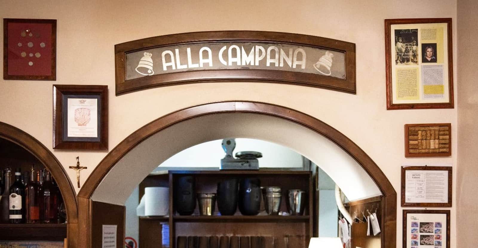 Arco in legno con insegna 'Alla Campana' sopra un passaggio verso il retro locale, cornici alle pareti.