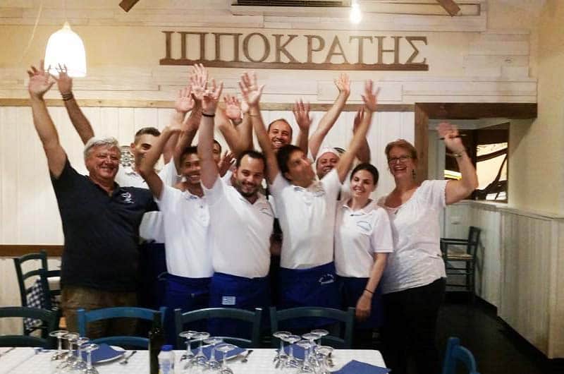 Gruppo di camerieri in polo bianca alza le braccia festante sotto una scritta greca a parete.