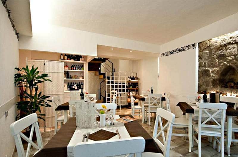 Sala da pranzo con tavoli in legno scuro apparecchiati, sedie bianche e piante verdi vicino a una libreria di bottiglie.