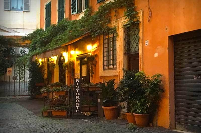 Facciata color ocra coperta di rampicanti, con tavoli illuminati sotto un pergolato e un'insegna nera con la scritta RISTORANTE.