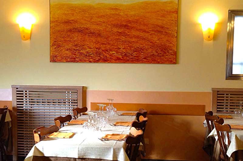 Sala con tavoli apparecchiati di bianco, sedie in legno e un grande quadro color ocra tra due applique.