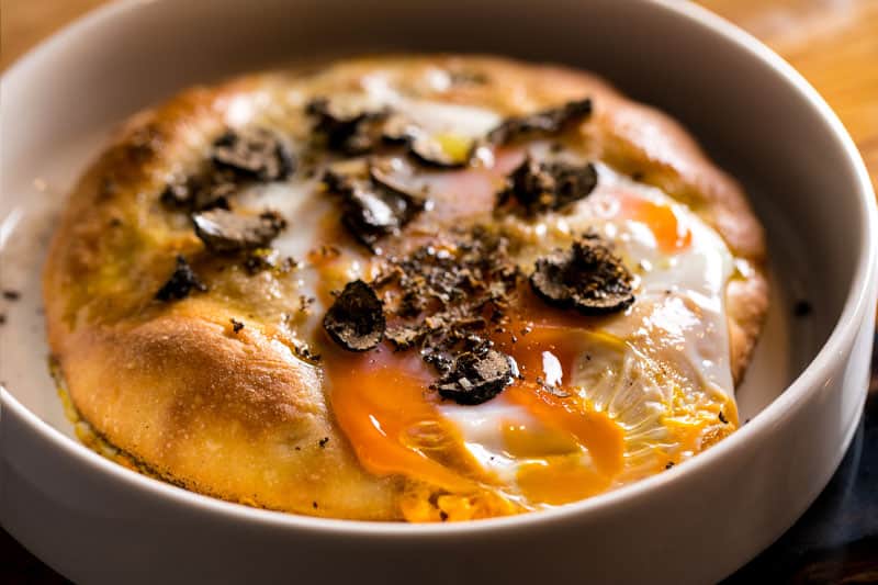 Focaccia tonda con uovo al centro e lamelle di tartufo nero, servita in una ciotola bianca.