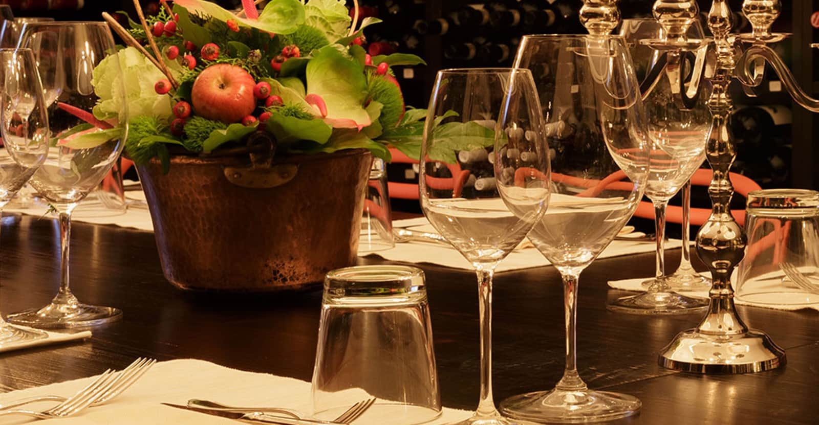 Bicchieri da vino vuoti e un candelabro in argento su un tavolo scuro, con un vaso di rame pieno di foglie e bacche rosse.