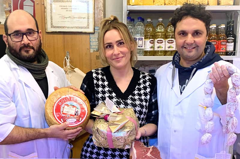 Due persone in camice bianco e una donna al centro, con una forma di formaggio, un pacco dorato e salumi appesi.