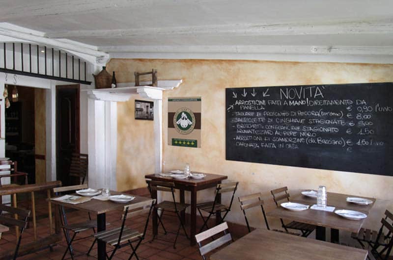 Sala con tavoli in legno apparecchiati e una grande lavagna nera con scritte in gesso bianco sulla parete.