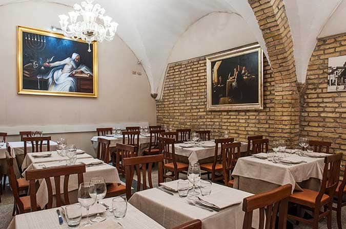 Sala da ristorante con soffitto a volta, pareti in mattoni e quadri in cornice dorata, tavoli apparecchiati con tovaglie chiare.