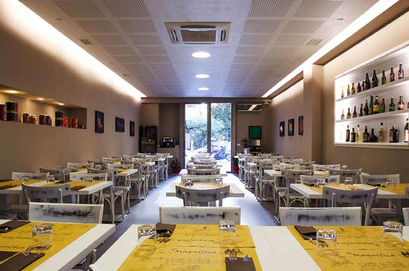 Sala di ristorante con tavoli dalle tovaglie gialle, sedie bianche e scaffali di bottiglie illuminati sul fondo.