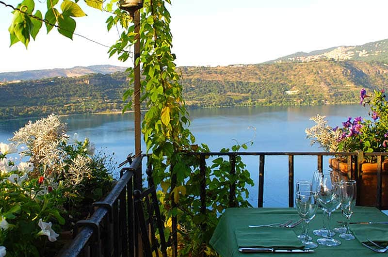 Tavolo apparecchiato con tovaglia verde su una terrazza affacciata su un lago circondato da colline.